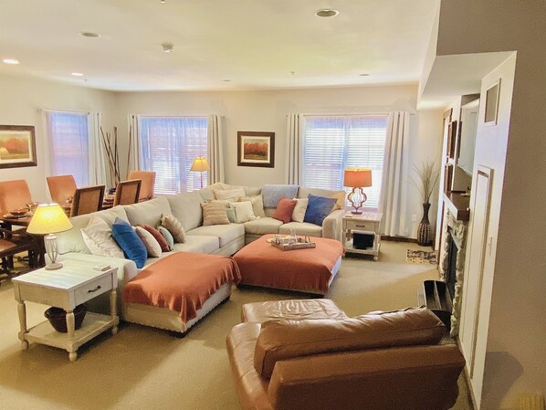 A spacious living room with a large sofa, a coffee table, and a side table with a lamp. There are two windows with curtains, a painting on the wall, and a fireplace. - Soaring Eagle 301 Ski In Out! (Snowshoe)
