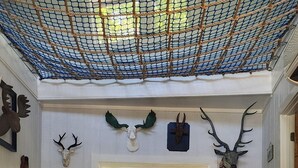 A cozy room with a ceiling net, a green sofa, a dining table with chairs, and wall-mounted deer antlers. - 2-story treehouse with a cargo net connecting the upstairs lofts, a large balcony with a slide to the ground level, sleeps 10. (Mill Spring)