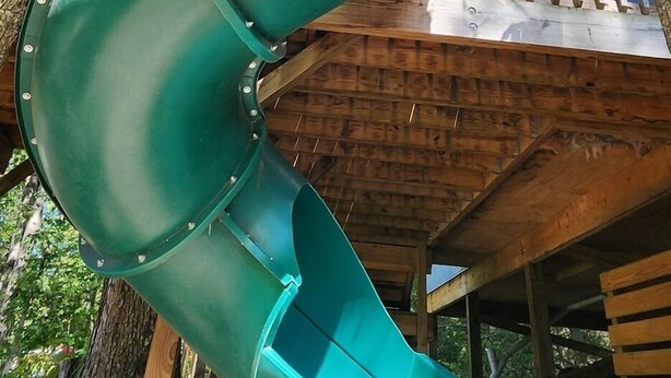 A green slide in a wooded area with a wooden deck and a house in the background. - 2-story treehouse with a cargo net connecting the upstairs lofts, a large balcony with a slide to the ground level, sleeps 10. (Mill Spring)
