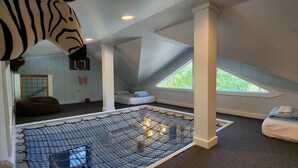A room with a sloped ceiling, a netted play area, a bean bag, a bed, and a window with a view of greenery. - 2-story treehouse with a cargo net connecting the upstairs lofts, a large balcony with a slide to the ground level, sleeps 10. (Mill Spring)