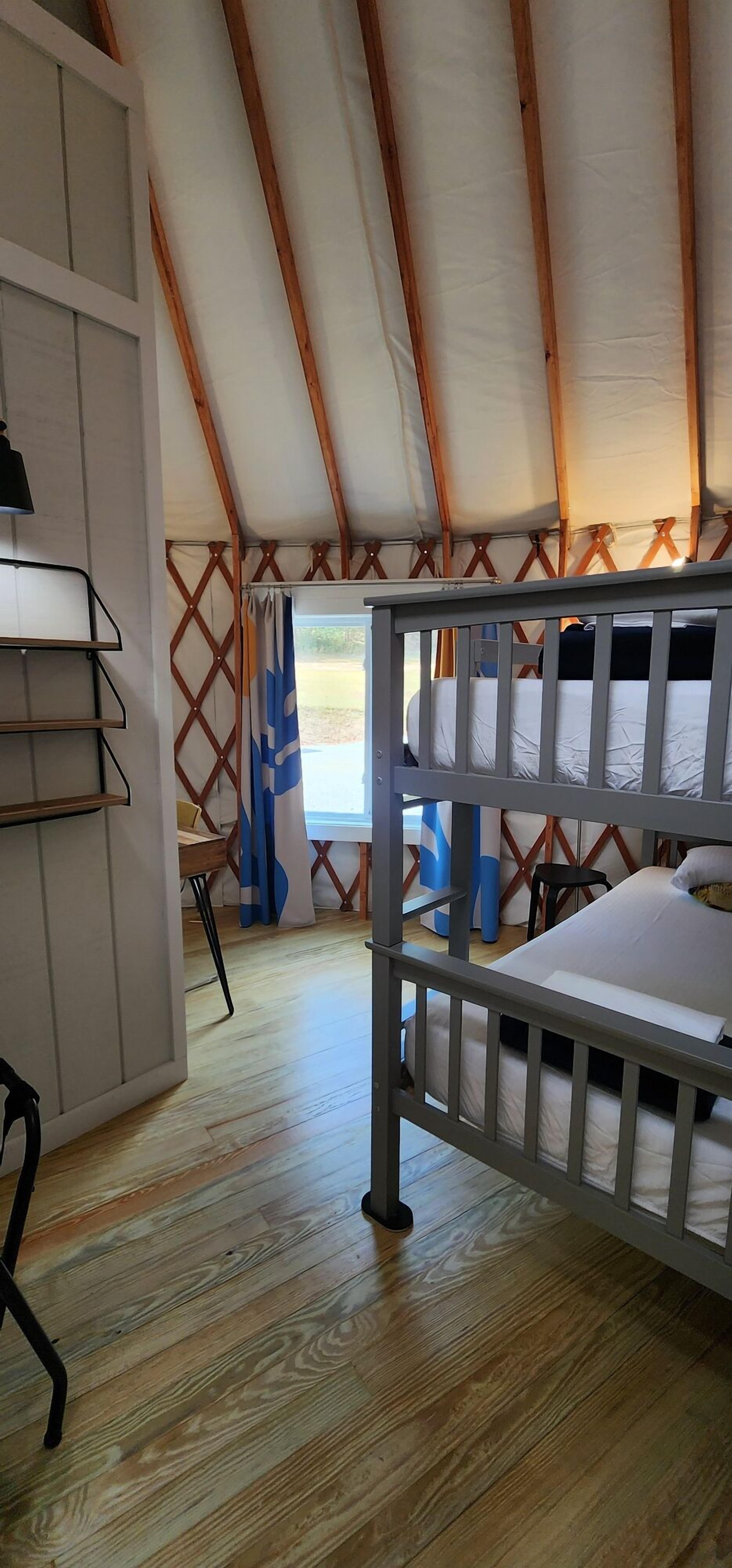 A room with a bunk bed, wooden flooring, a desk with shelves, and a window with curtains.