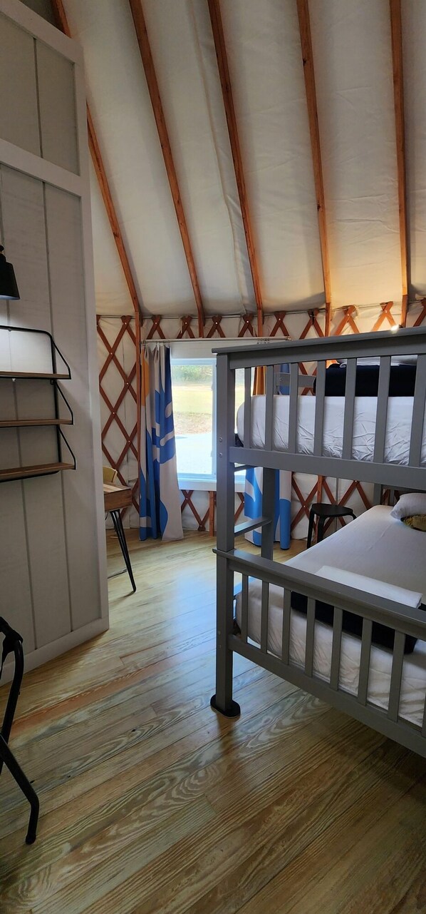Een kamer met een stapelbed, houten vloer, een bureau met planken en een raam met gordijnen.