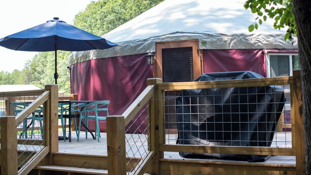A yurt with a covered porch, a grill, and a wooden fence. - 30' Yurt with a large deck, on the creek, full kitchen, dome skylight, 2 bedrooms, sleeps 5. (Mill Spring)