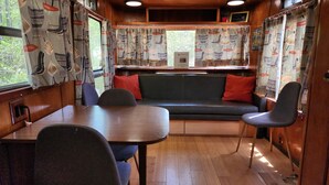 A cozy RV interior with a dining table, two chairs, a sofa with red cushions, and patterned curtains. - Vintage 1955 Imperial Mansion, deck on the creek, easy access to pool and playground, sleeps 5. (Mill Spring)
