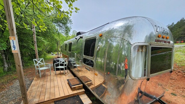 Een klassieke caravan met een houten terras, twee witte stoelen en een tafeltje.