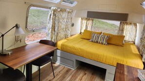 Een kleine slaapkamer met een bed, een bureau en een stoel. Er zijn ramen met gordijnen, een plafondventilator en uitzicht op de omgeving.