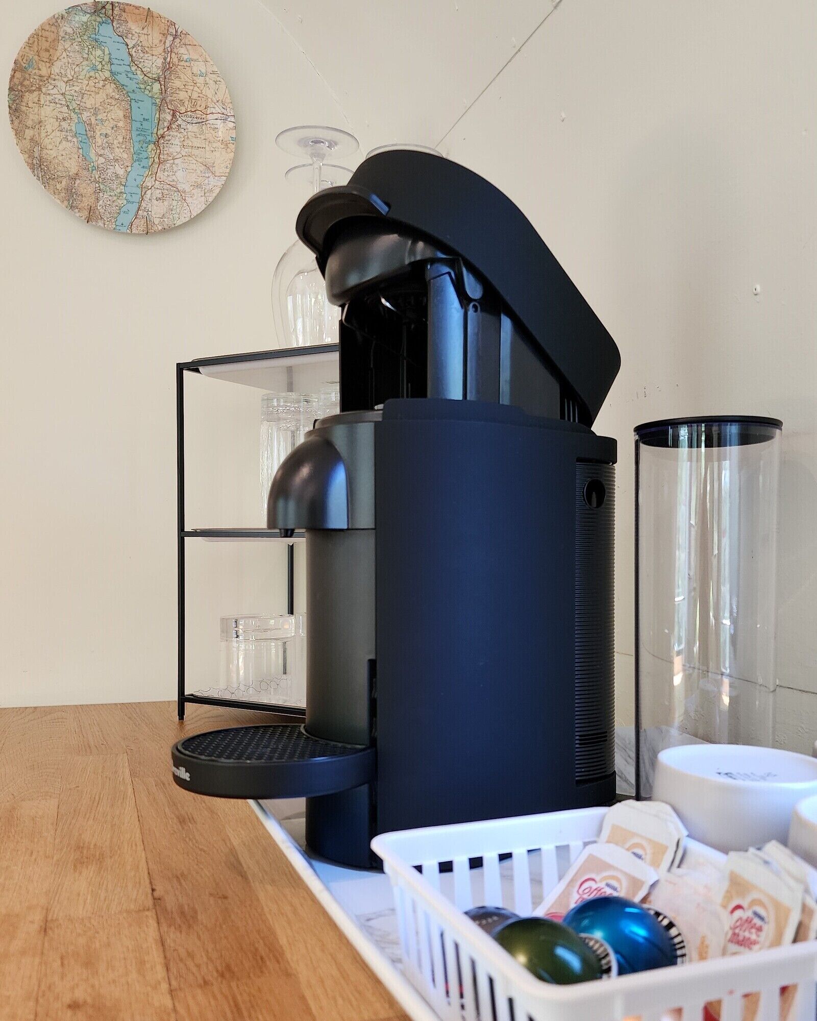 A coffee machine with a black base and a transparent water tank, a glass coffee pot, and a basket of coffee pods on a wooden table.
