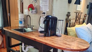 A compact kitchen with a wooden countertop, a sink, and a coffee machine. There is a shelf above the sink holding various items, and a wall-mounted lamp to the right. - 16' Yurt, Dog Friendly, Deck, Dome Skylight, Sleeps 2. (Mill Spring)