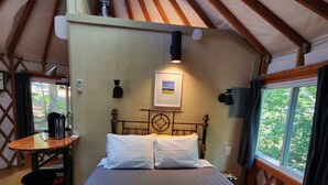 A cozy, circular room with a bed, a small table, and a chair. There is a window with curtains and a view of greenery outside. - 16' Yurt, Dog Friendly, Deck, Dome Skylight, Sleeps 2. (Mill Spring)