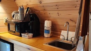 Et kompakt køkken med trævægge og en træbordplade. Det indeholder en vask, elkedel, kaffemaskine og en kurv med service.