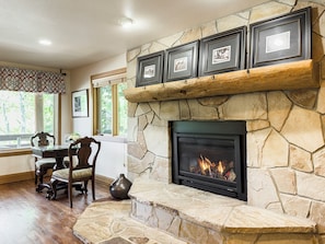 A stone fireplace with framed pictures above, a dining area with a table and chairs, and a window with a view of trees. - In Lower Deer Valley and less than 2 miles to Main Street! (Park City)
