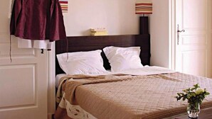 A neatly made bed with white pillows and a beige quilt, a wooden nightstand with a vase of flowers, and a hanging garment on a wooden hanger. - Entre mer & campagne | Maison T3 + accès piscine (Roquebrune sur Argens)