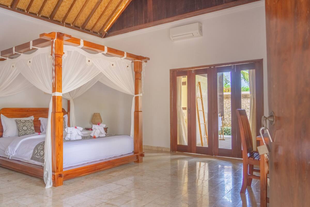 Una cama con dosel y cuatro columnas con ropa blanca, cabecera de madera y mesita de noche con lámpara.