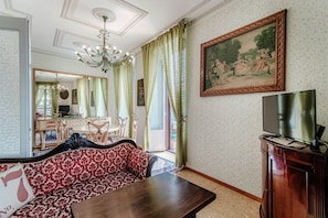 Een woonkamer met een bank, een tafel, een televisie, een schilderij en een eethoek met stoelen en een tafel.