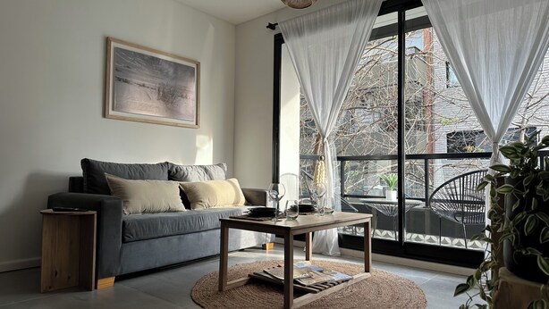 Una sala de estar moderna con un sofá gris, una mesa de centro de madera y un ventanal con cortinas ligeras.