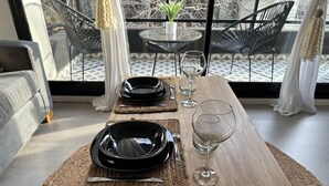 Una mesa de comedor dispuesta con platos negros, cubiertos y una copa de vino, rodeada de individuales tejidos y una superficie de madera.