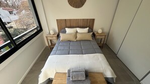 Un dormitorio moderno con una cama grande, cabecera de madera, dos mesitas de noche, un banco y vistas al exterior.