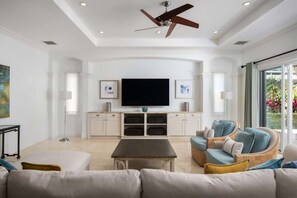A modern living room with a ceiling fan, a flat-screen TV, wicker furniture, and a large window.
