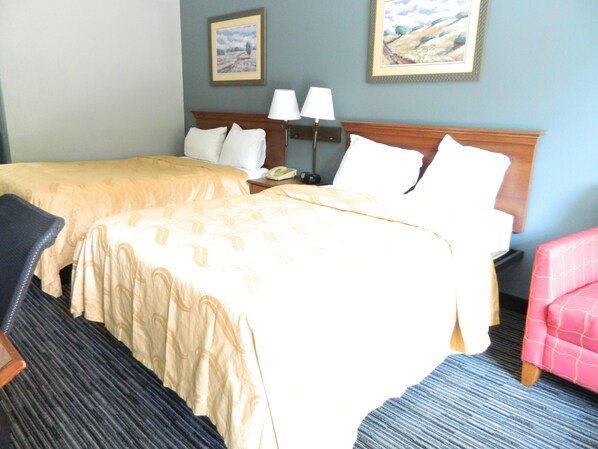A hotel room with two beds, a nightstand with a lamp, a chair, and two framed pictures on the wall. - Travel Inn Atlanta (Atlanta)