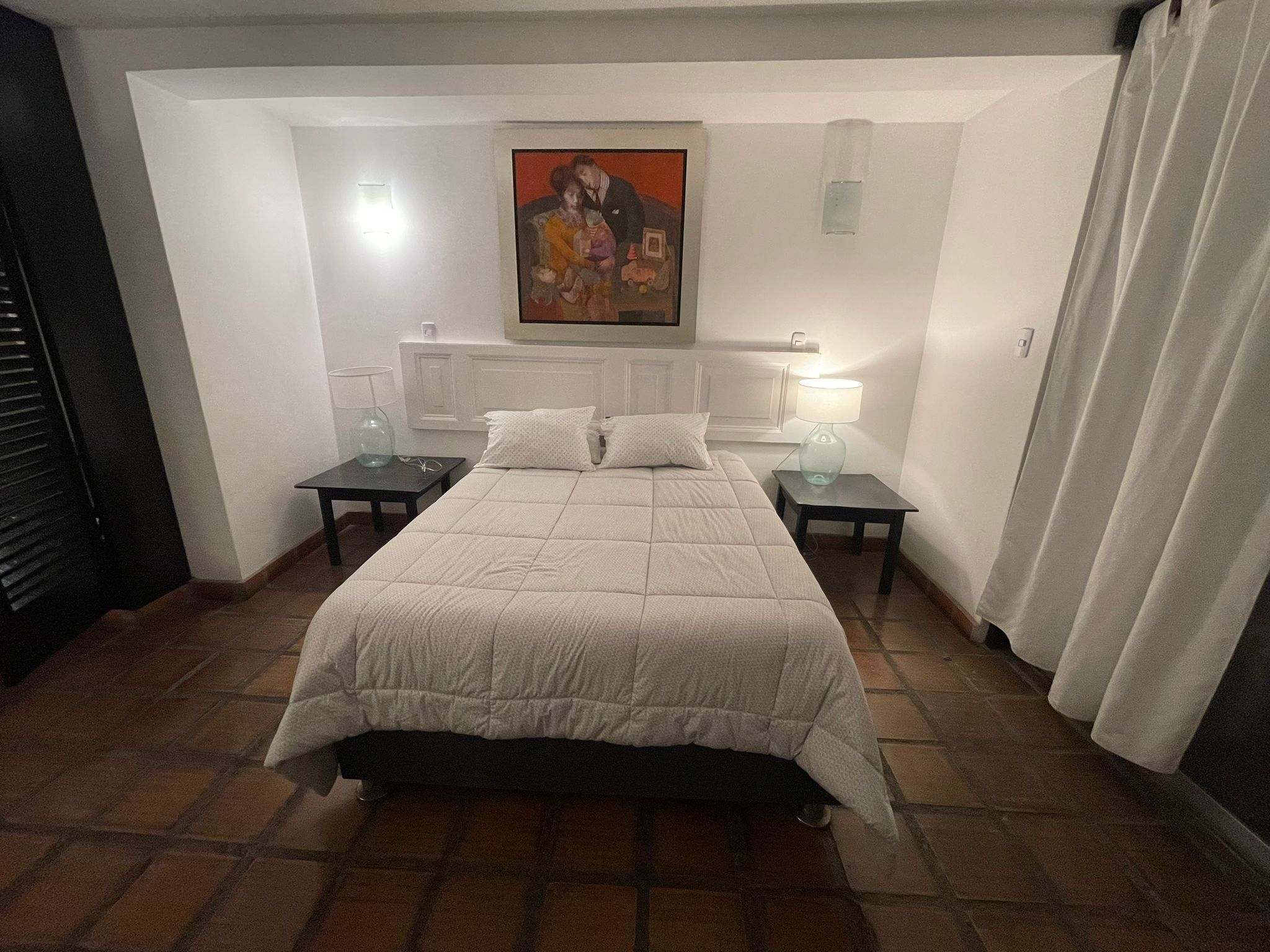 Un dormitorio con una cama, dos mesitas de noche, un aparador, un cuadro en la pared y una ventana con cortinas.
