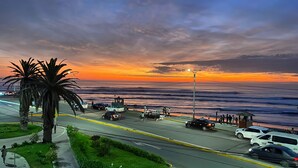 A coastal road with parked cars, a palm tree, and a sunset over the ocean.