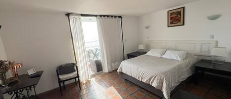 Habitación de hotel con cama, escritorio, silla, ventana con cortinas y un pequeño balcón.