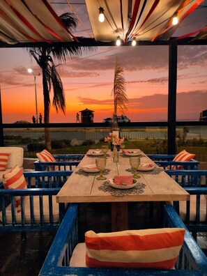Un comedor con vistas al atardecer, una mesa de madera dispuesta con platos y vasos, y cojines a rayas azules y rojas.