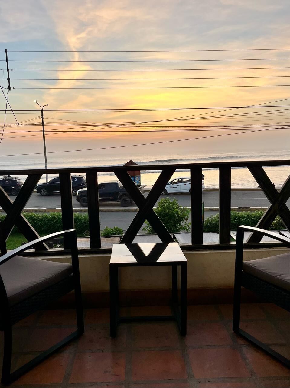 A balcony with a table and chairs, overlooking a street with parked cars and a body of water in the background.