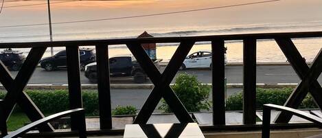 A balcony with a table and chairs, overlooking a street with parked cars and a body of water in the background.