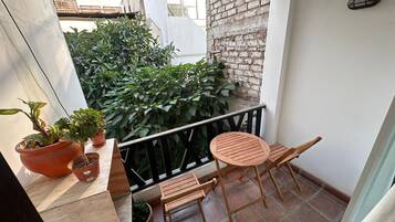 A small balcony with a wooden table and chairs, potted plants, and a view of a brick wall and greenery.