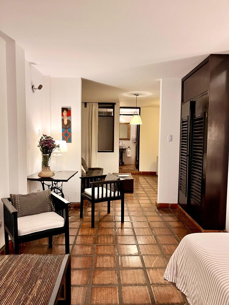 A hotel room with a bed, a small table with a vase of flowers, a sofa, and a chair. There is a bathroom visible in the background.