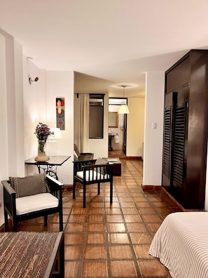 Habitación de hotel con cama, una pequeña mesa con un florero, un sofá y una silla. Se ve un baño al fondo.