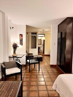 Habitación de hotel con cama, una pequeña mesa con un florero, un sofá y una silla. Se ve un baño al fondo.