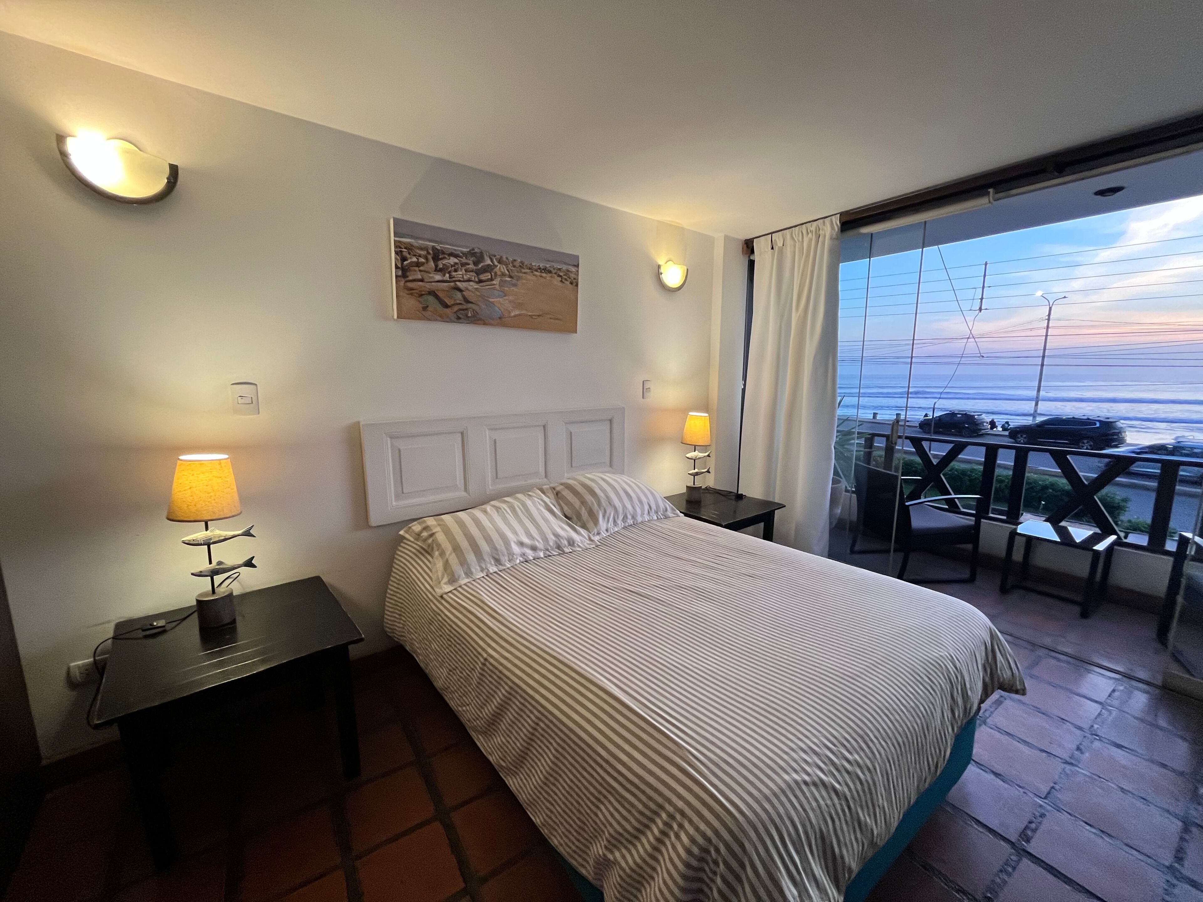 A hotel room with a bed, bedside lamps, a painting, and a balcony with a view.
