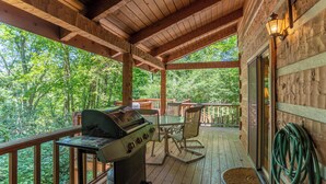 A wooden deck with a grill, a table, and chairs, overlooking a forested area. - Hummingbird Hollow (Banner Elk)