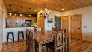 A wooden dining table with chairs, a chandelier, and a kitchen area with wooden cabinets and a microwave. - Sugar Haven (Banner Elk)