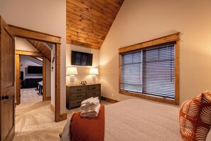 A bedroom with a wooden ceiling, a large window with blinds, a bed with a red blanket, a dresser with lamps, and a TV on the wall. - Price Peak at Eagles Nest (Banner Elk)