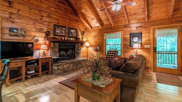 A cozy cabin interior with a stone fireplace, wooden walls, a ceiling fan, and a television. - Trillium (Banner Elk)