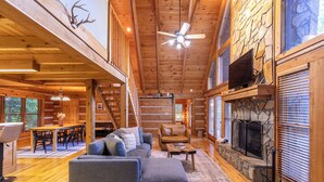 A spacious cabin interior with a large sofa, a fireplace, a dining area, and a staircase leading to an upper level. - Highlander Cabin (Deep Gap)
