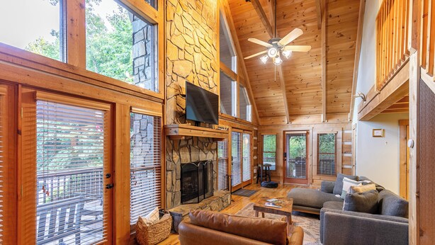 A spacious living room with a stone fireplace, wooden ceiling, and large windows. - Highlander Cabin (Deep Gap)