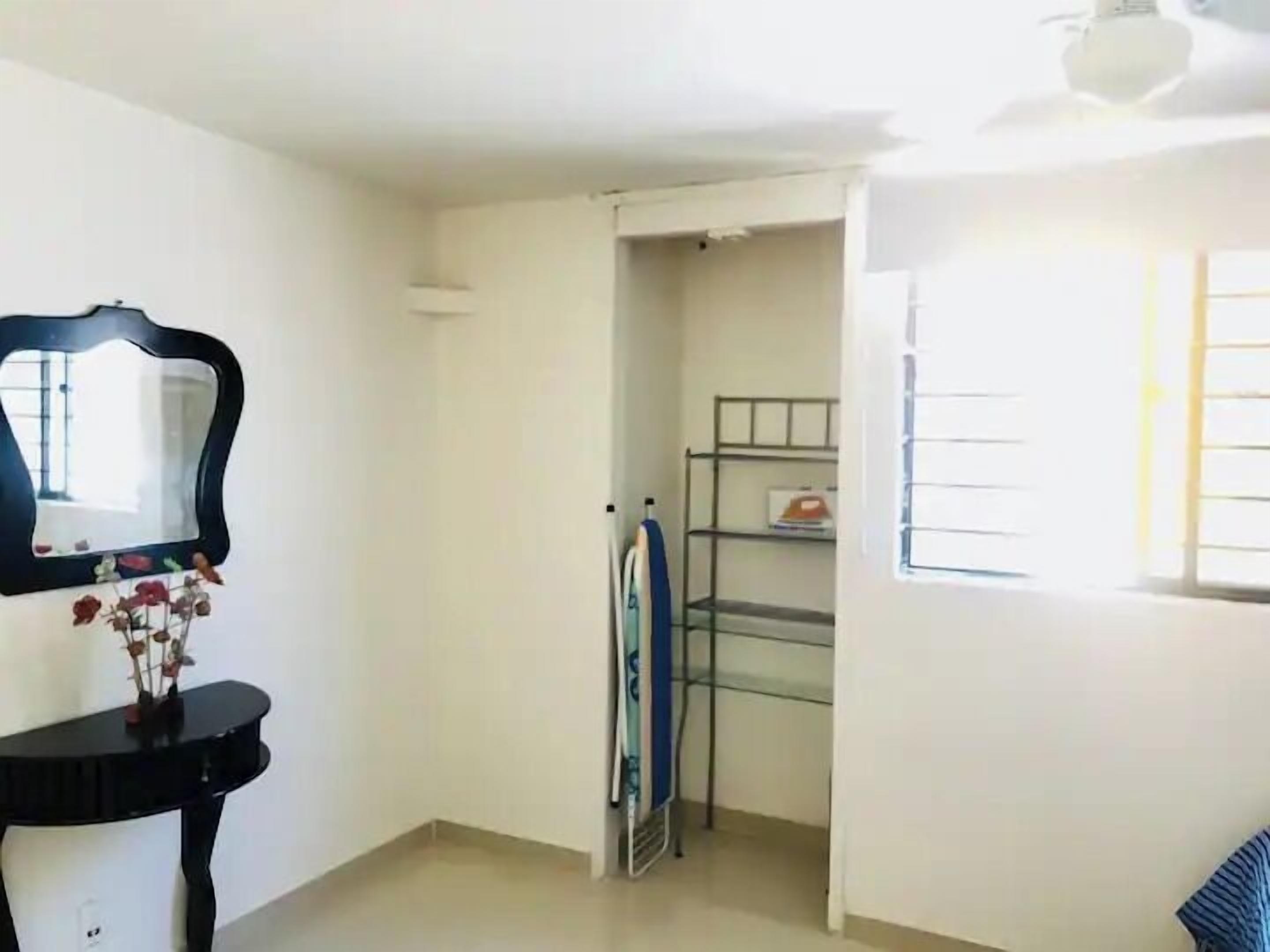 A room with a mirror, a small table with flowers, a metal shelf unit, and a window with blinds.