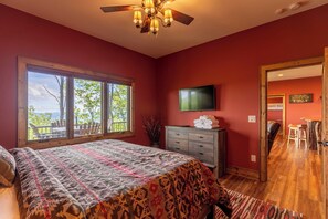 A bedroom with a large bed, a ceiling fan, a TV, and a window with a view of trees. - The Refuge at Eagles Nest (Banner Elk)