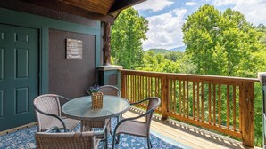 A covered outdoor seating area with a table and chairs, a wooden railing, and a view of green trees and hills. - Still Lake Haven at Echota (Boone)