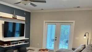 A modern living room with a large gray sectional sofa, a flat-screen TV on a white wall, a ceiling fan, and a door with glass panels. - Crow Creek 1706 | Course-Ready, Comfort (Calabash)