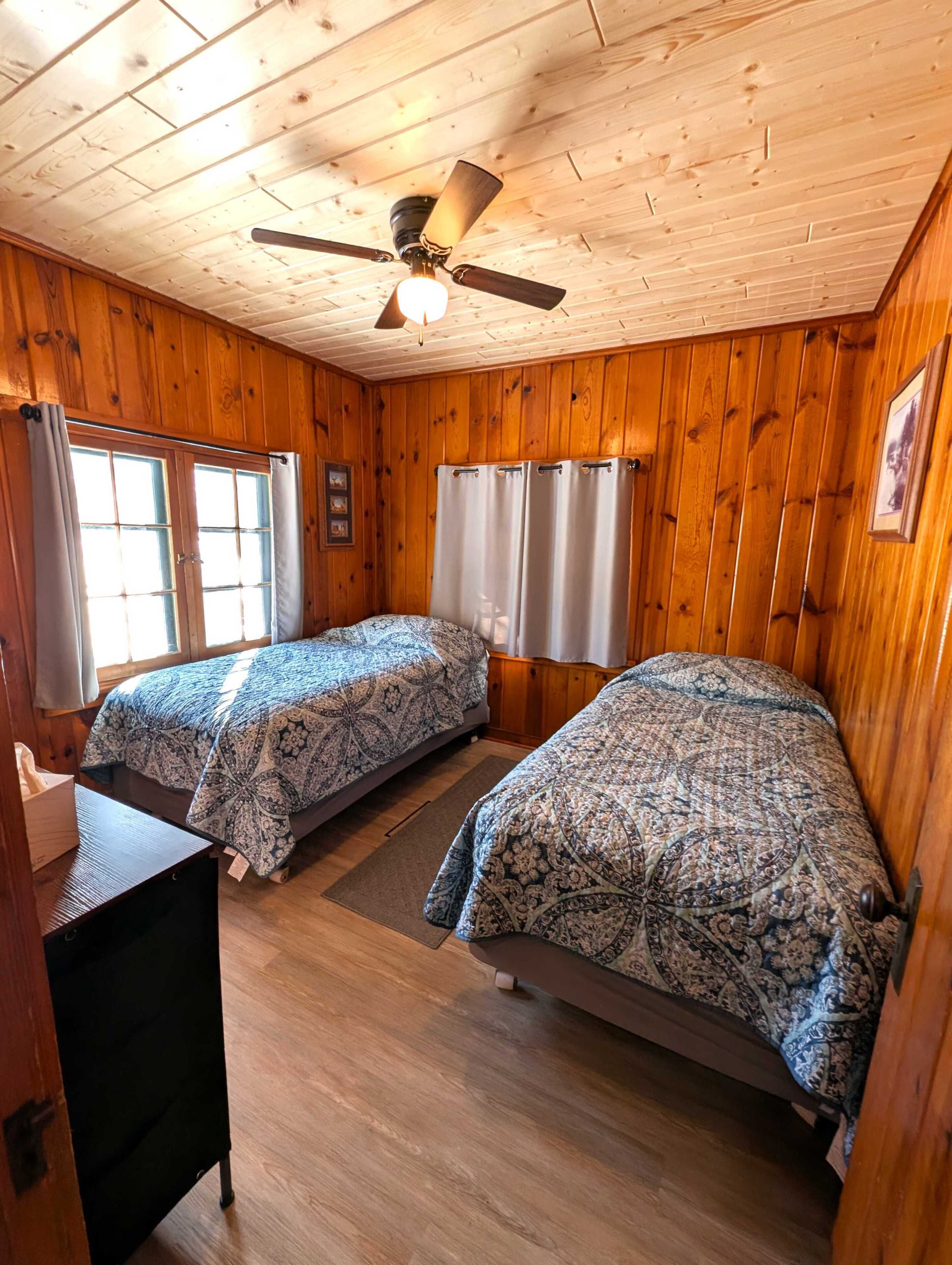 A cabin with two beds, wooden walls, a ceiling fan, and a window with curtains.
