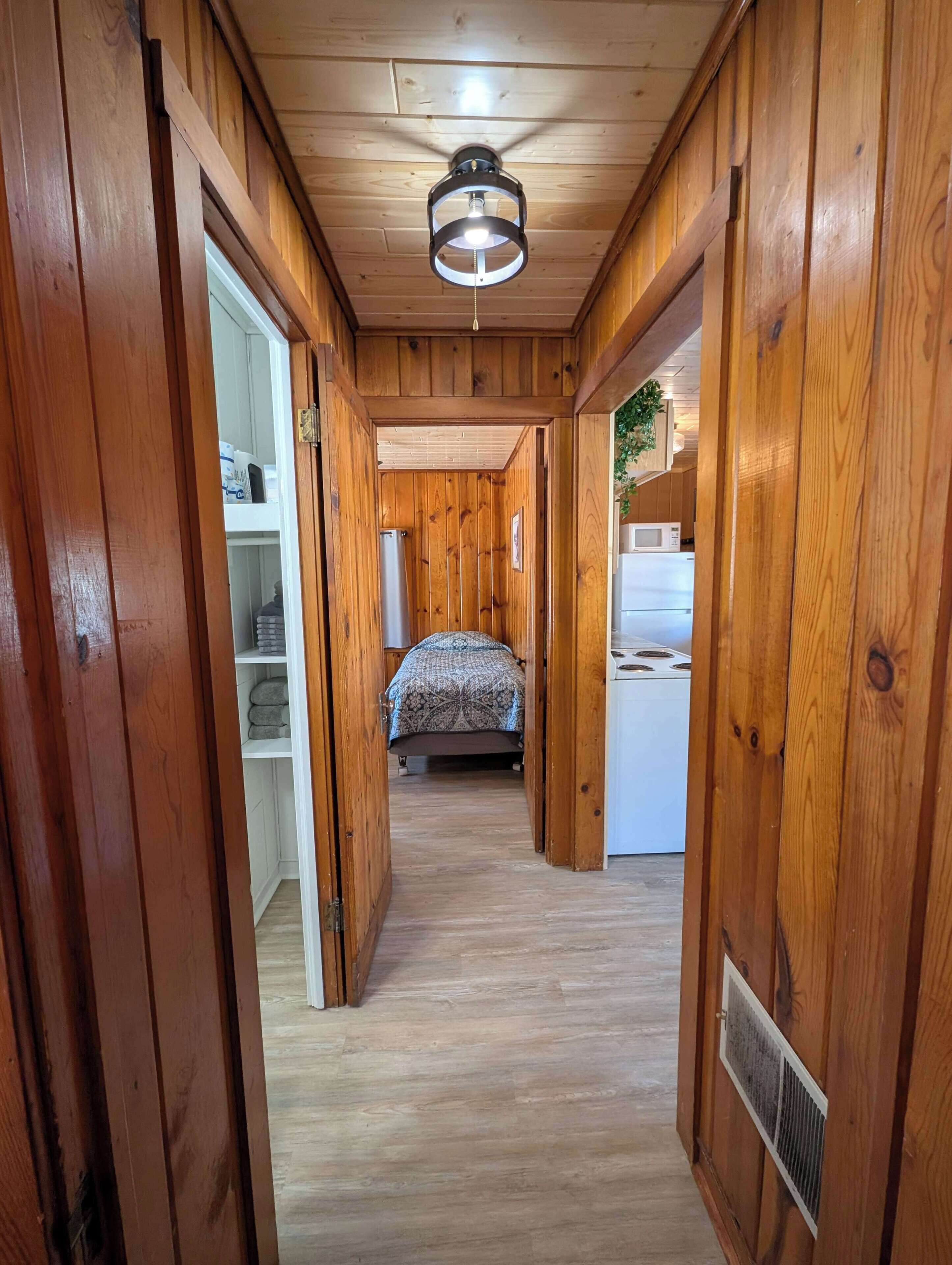 Ein heller Holzboden-Flur führt zu einem Schlafzimmer mit einem Bett, einem weißen Schrank und einer Deckenleuchte.