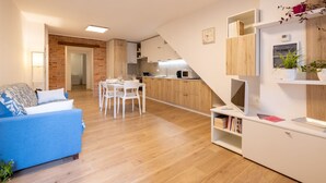 A modern living room with a blue sofa, a dining area with white chairs, and a kitchen with wooden cabinets.