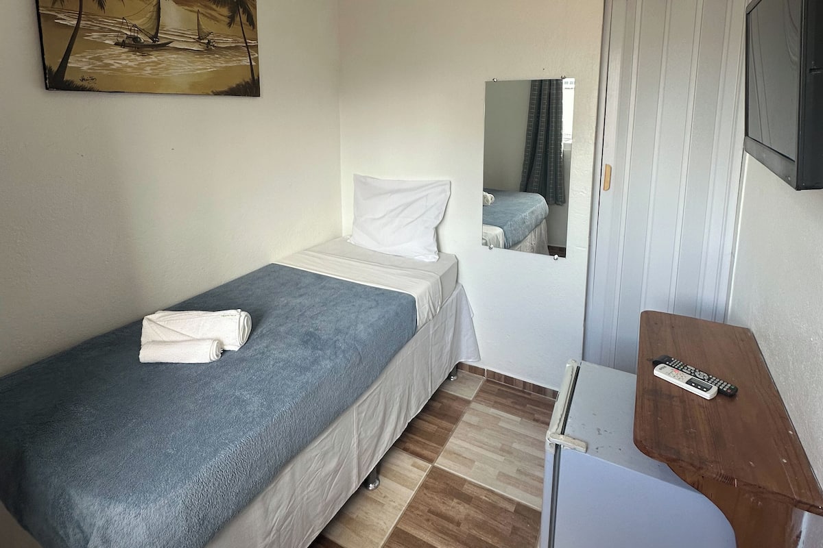 A small, neatly arranged room with a single bed, a small table, a TV, and a painting on the wall.