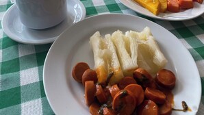A table with a checkered tablecloth, a cup of coffee, and plates of food including sausages, potatoes, and a slice of cake. - Pousada Porto Navegantes (Ipojuca)