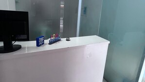 A reception desk with a glass partition, a computer monitor, and a small blue box with text. - Pousada Porto Navegantes (Ipojuca)